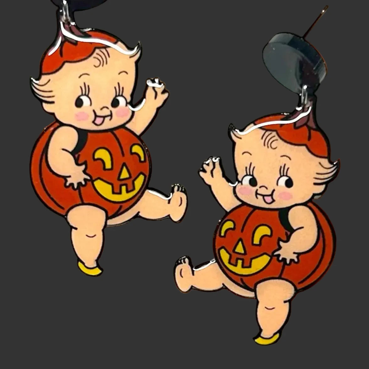Two Kewpie vintage-style enamel pins shaped like mischievous children in red hats and pumpkin costumes, shown against a blurred background of pumpkins.