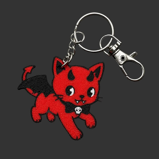 A red 3D printed kewpie devil cat keychain with black wings, a black bow tie, and a silver clasp, displayed on a round black slate coaster.