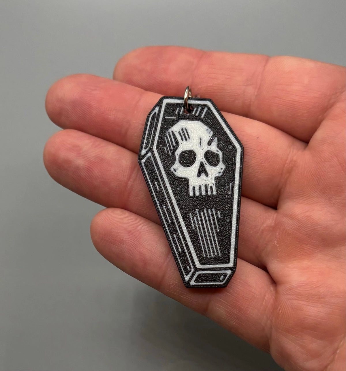 A small coffin-shaped pendant or charm with a white skull graphic on a black background, held in a person's hand against a plain gray backdrop.