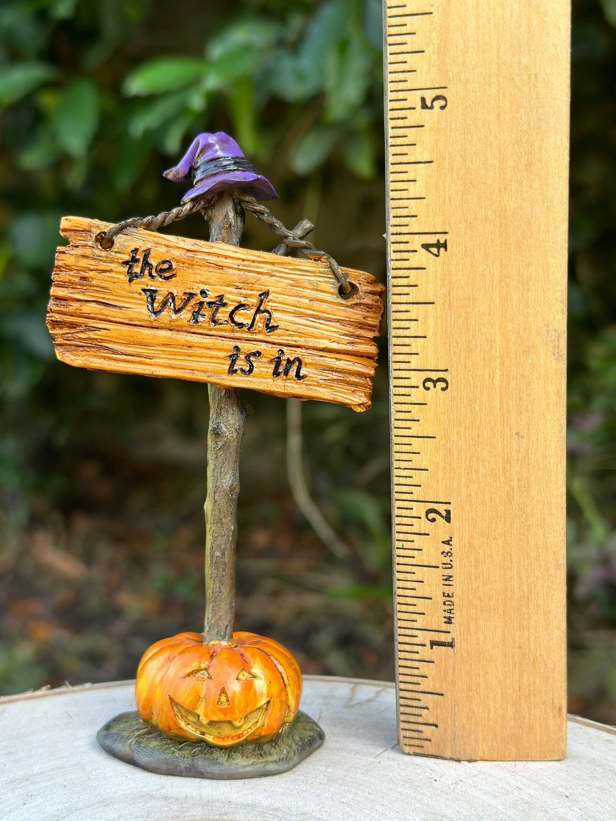 Size reference photo of The Witch Is In sign miniature next to a ruler showing approximately 4.5 inches tall, with purple witch hat and jack-o-lantern base