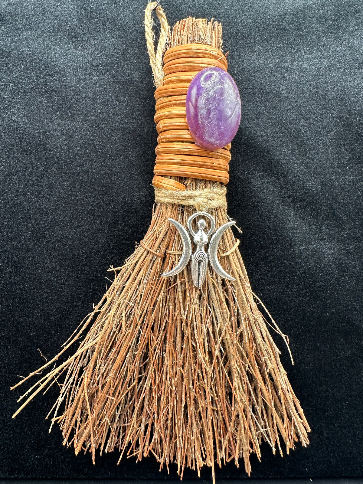 Handcrafted Triple Moon Goddess witch besom with natural raffia fibers, wrapped cord, polished amethyst crystal, and silver crescent moon charm on a dark background