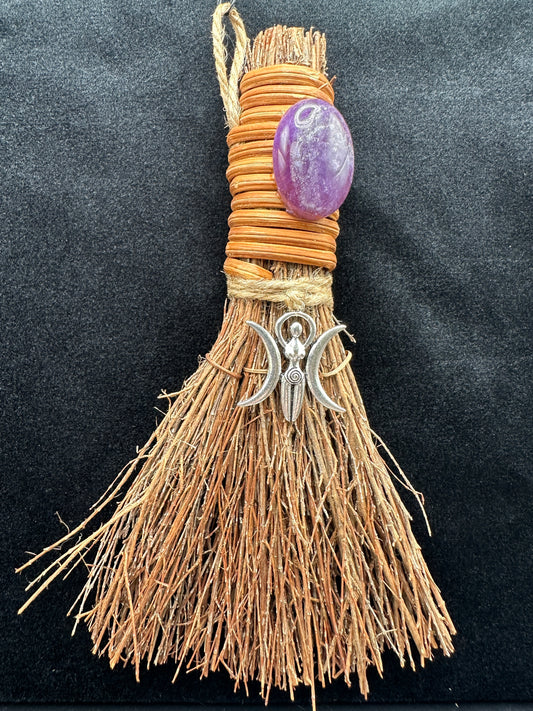 Handcrafted Triple Moon Goddess witch besom with natural raffia fibers, wrapped cord, polished amethyst crystal, and silver crescent moon charm on a dark background