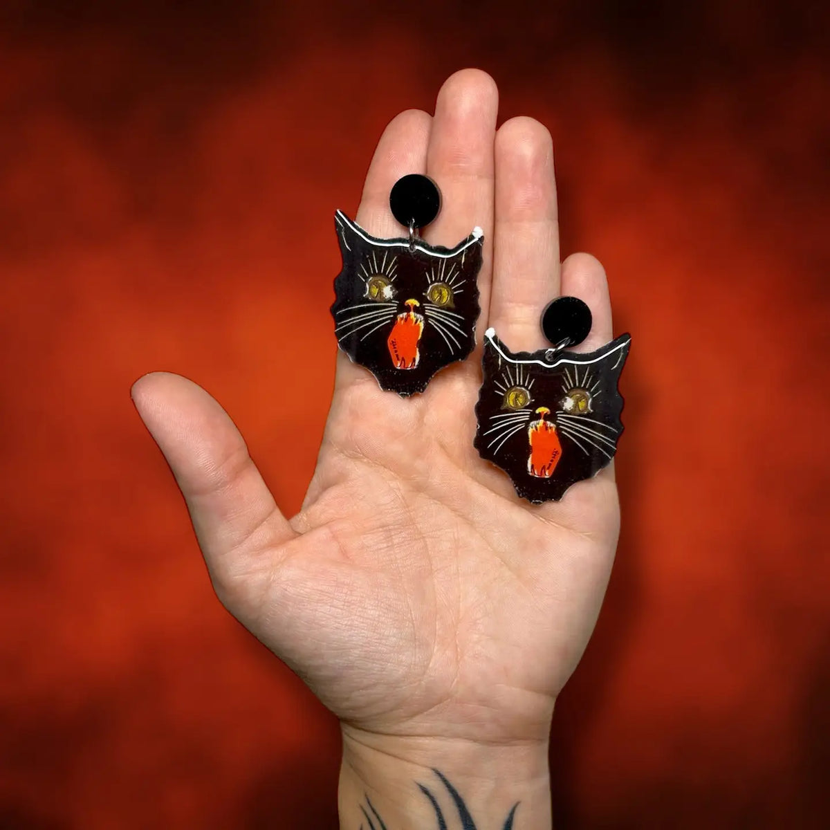 A hand holding a pair of black cat-shaped earrings with gold and red enamel details against a textured red background.