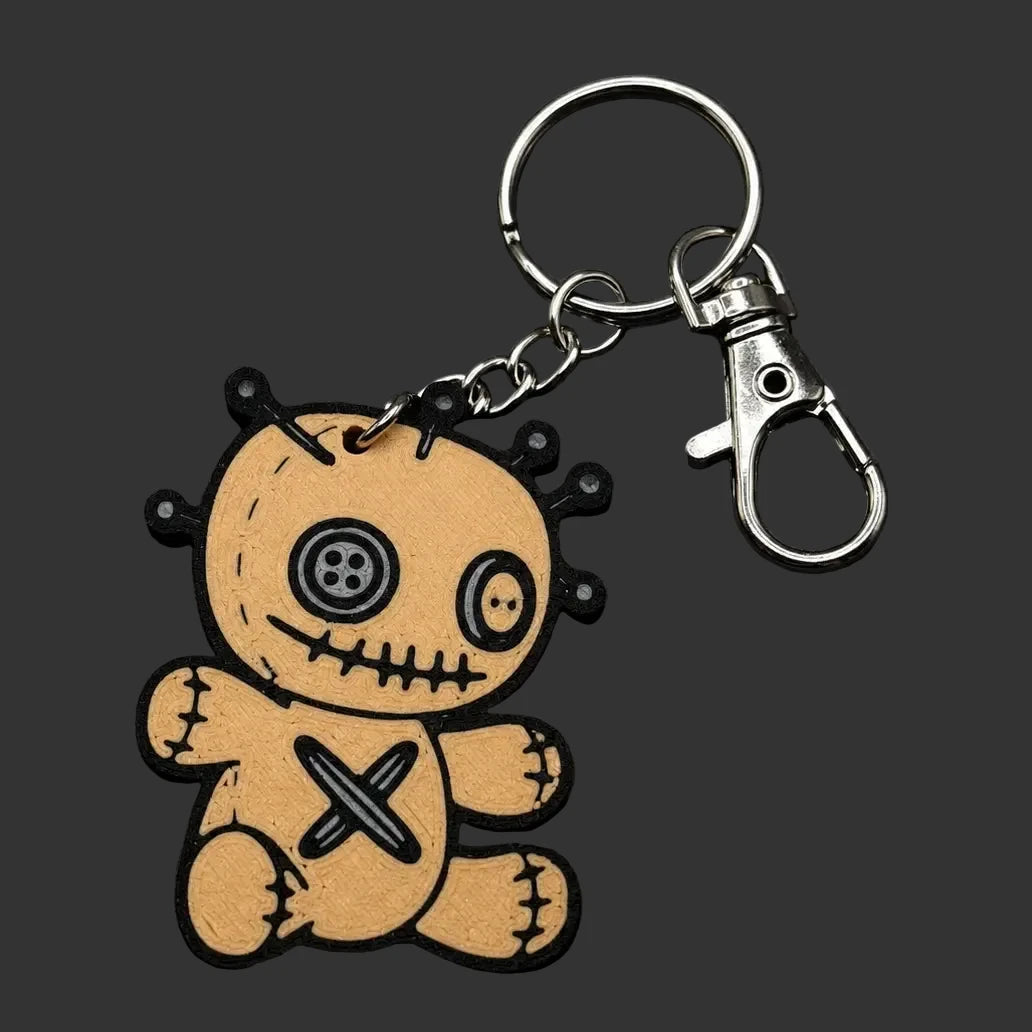 A voodoo doll keychain featuring a tan vinyl figure with black stitched details, a button eye, and a cross on the chest, attached to a silver metal keyring and lobster clasp, displayed on a dark slate background.