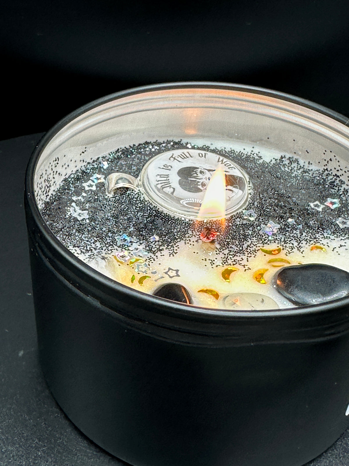 A black metal candle tin with a clear clear wax decorated in black glitter and stars with black and gold crystals lit with a small flame. The candle wax is white and filled with black glitter, small star-shaped embellishments, and gold crescent moon details. A Wednesday Adam's removable pendant is in the candle.