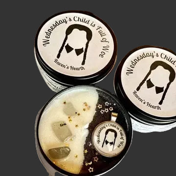 Three round tin candles with black lids and white labels featuring a black-and-white portrait of Wednesday Addams and the text 'Wednesday's Child is Full of Life' and 'Haven's Hearth'. One tin is open, showing a cream-colored wax surface with gold flecks, translucent stones, and a small pendant charm.