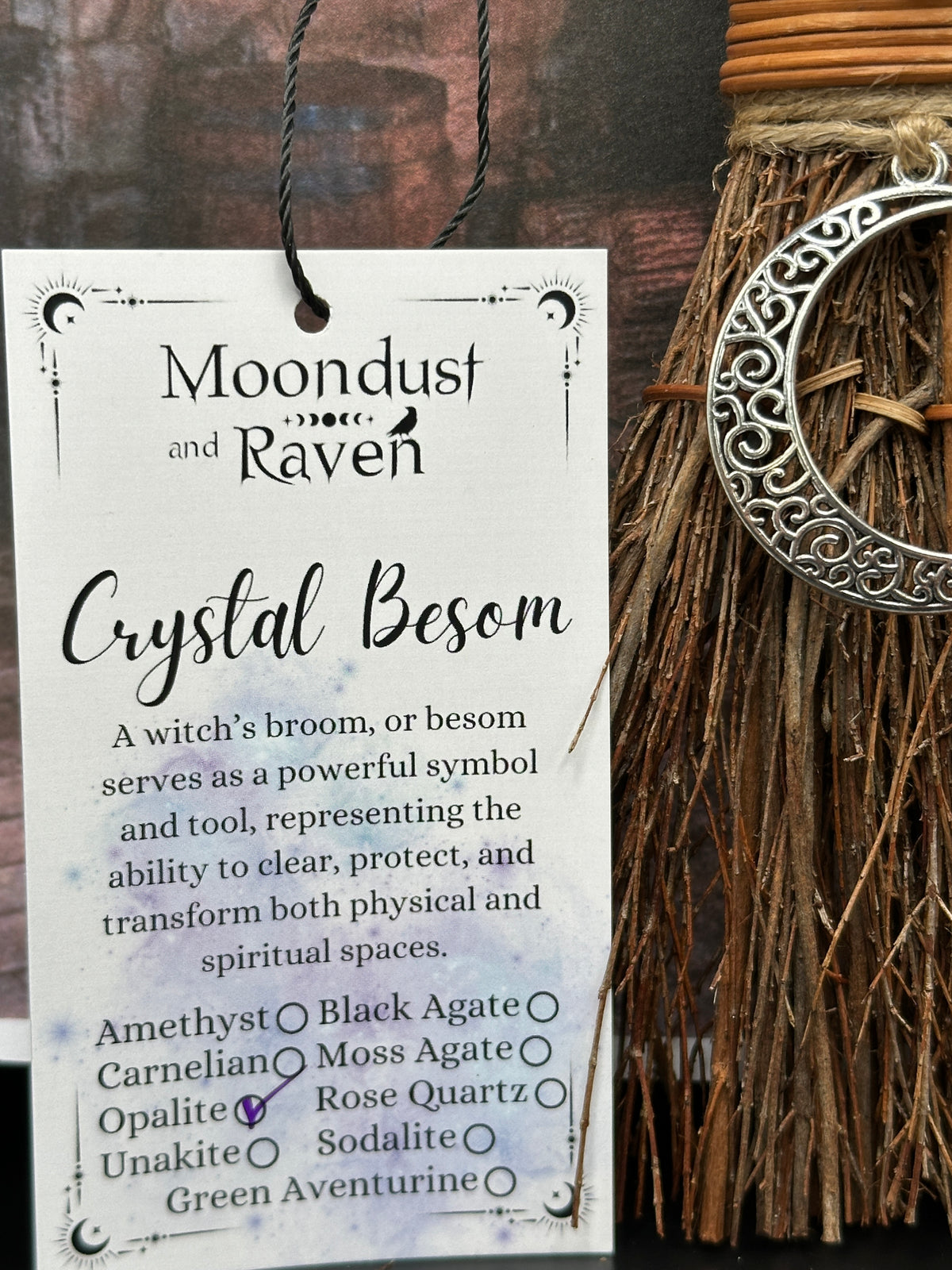 A product tag for 'Moondust and Raven Crystal Besom' is shown next to a handmade witch's broom with a silver crescent moon ornament, set against a rustic dark wood background.