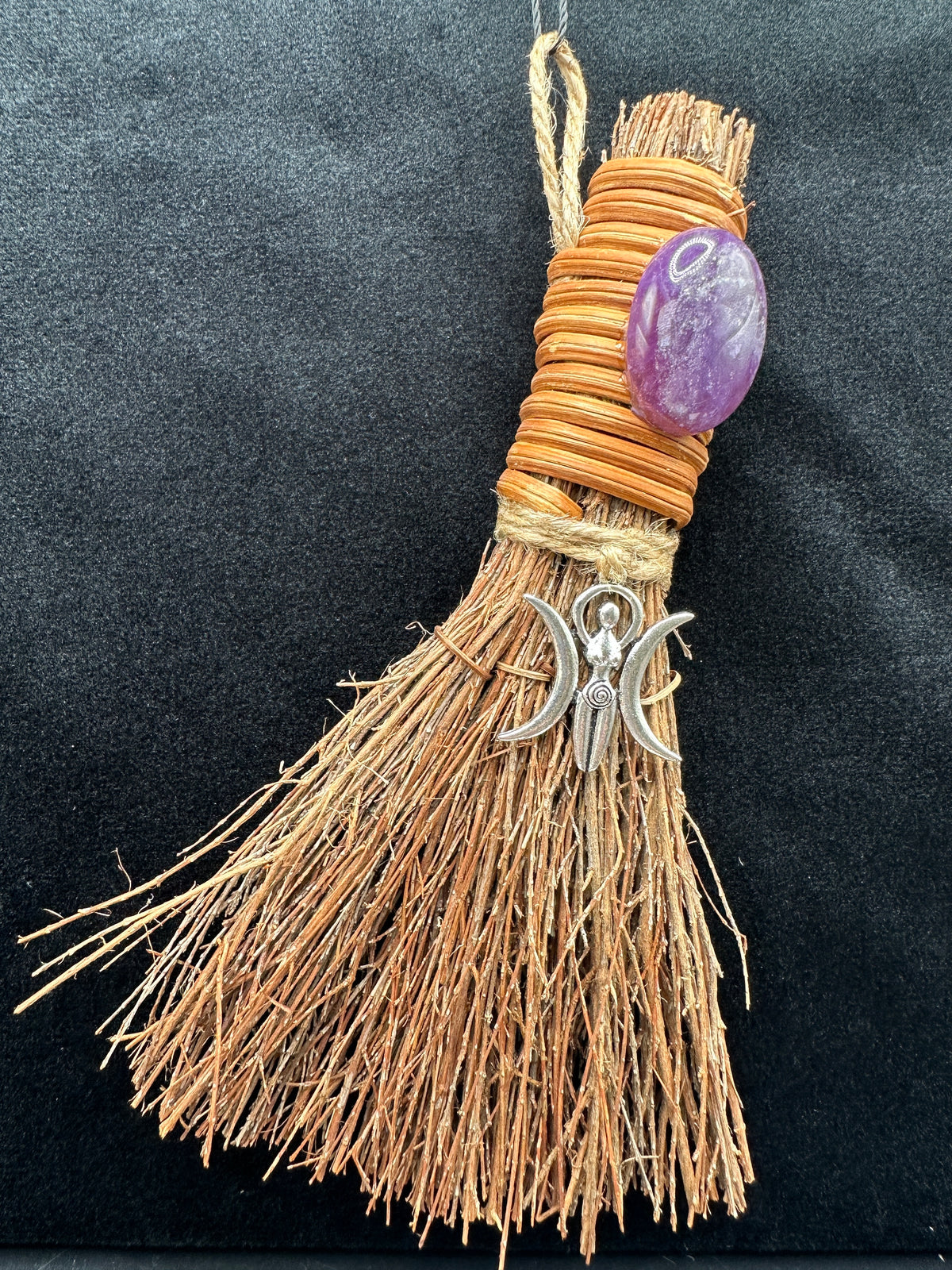 A handmade tassel witchy ornament featuring natural tan raffia fibers wrapped with light brown cord, adorned with a polished purple amethyst stone and a silver crescent moon and double moon charm, set against a dark textured background.
