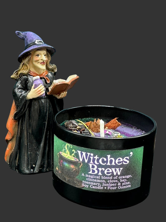 A miniature witch figurine in a black robe and purple hat stands beside a black tin candle labeled 'Witches' Brew' with a matching label. The candle contains visible herbs and wax pieces, and the background is a spooky blue-toned haunted-house scene.