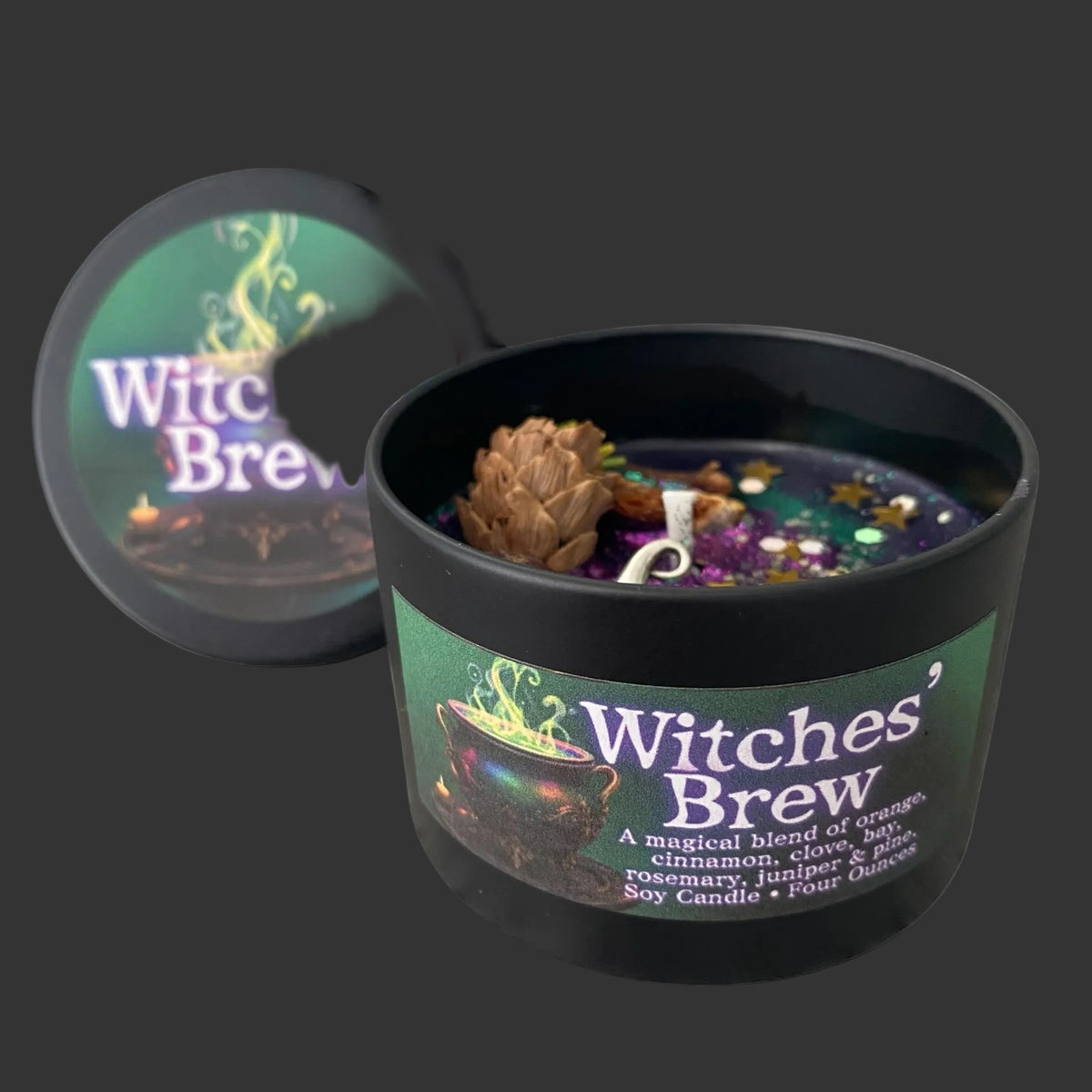 A black soy candle labeled 'Witches' Brew' with a matching lid leaning behind it on a white background. The open candle shows a dark wax surface with a pinecone, small dried botanicals, and a white wick.