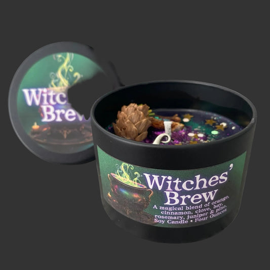 A black soy candle labeled 'Witches' Brew' with a matching lid leaning behind it on a white background. The open candle shows a dark wax surface with a pinecone, small dried botanicals, and a white wick.