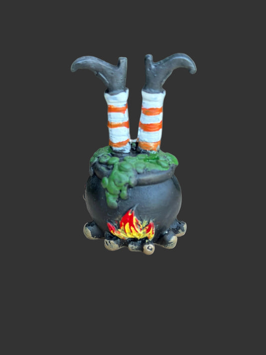A small Halloween decoration shaped like a black cauldron with green slime, a tiny red and yellow fire, and two witch legs with orange-and-white striped socks popping out, displayed on a round wooden slice with a blurred green garden background.