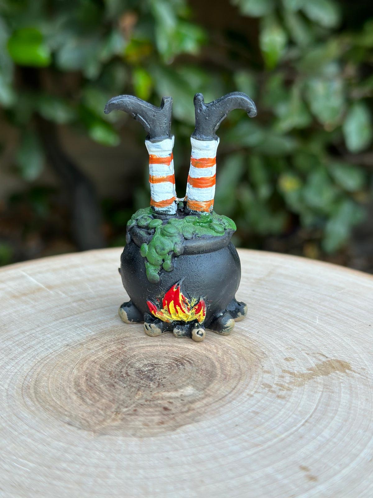 A small Halloween witch cauldron figurine sitting on a wooden surface with a blurred green garden background. The cauldron is dark gray with green slime, small red and yellow flames at the base, and two black-and-white striped legs sticking up from the top.