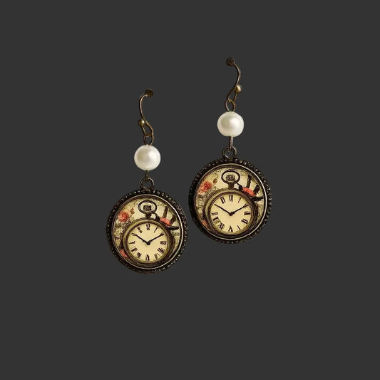A pair of steampunk-style drop earrings displayed on a white card. Each earring has an antique bronze hook, a white pearl bead, and a round pocket-watch pendant with a floral design and Roman numerals.