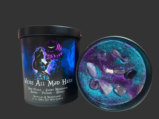 Candle with black tin  lid featuring an Alice in Wonderland character and text that says "we're all mad here", surrounded by decorative stones and flowers. The inside of the candle is decorated with purple and blue glitter and lavender crystals.