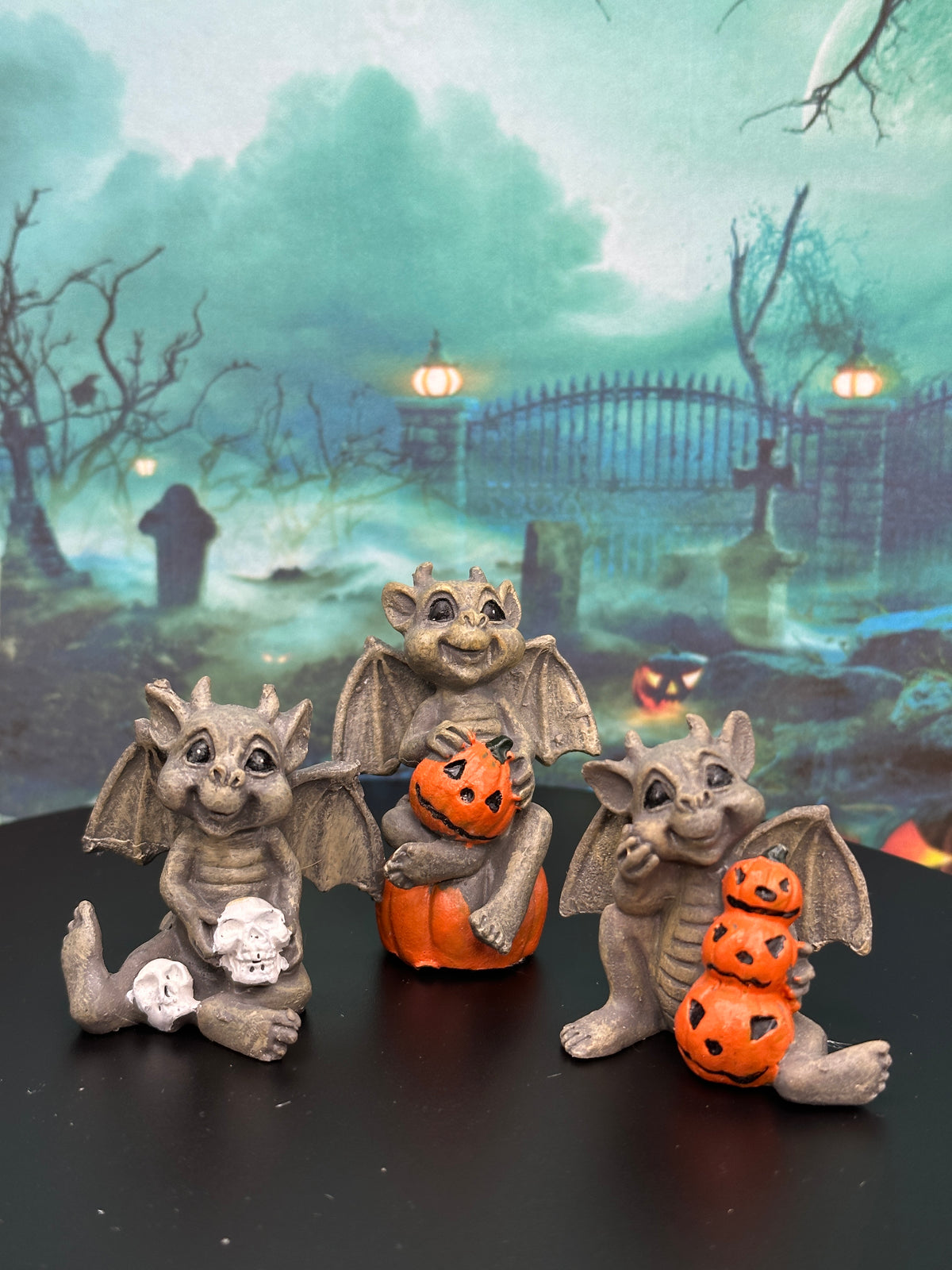 Three small gray stone-like gargoyle figurines posed on a black surface in front of a Halloween-themed backdrop showing a misty graveyard scene. The gargoyle on the left holds a white skull, the center gargoyle sits on an orange pumpkin, and the right gargoyle holds a stack of three orange jack-o'-lanterns.