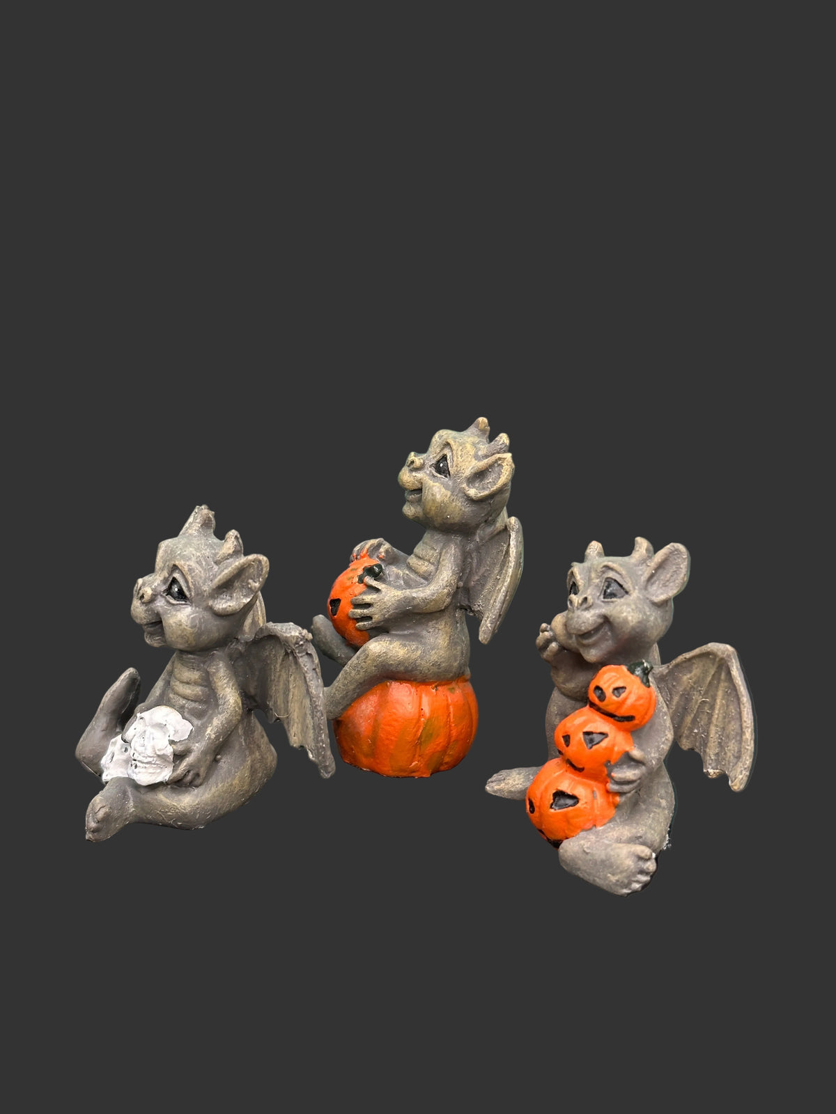Three miniature gray stone-like gargoyle figurines posed on a black surface. One gargoyle sits on an orange pumpkin, and another holds a stack of orange jack-o'-lanterns; a third holds a white skull.