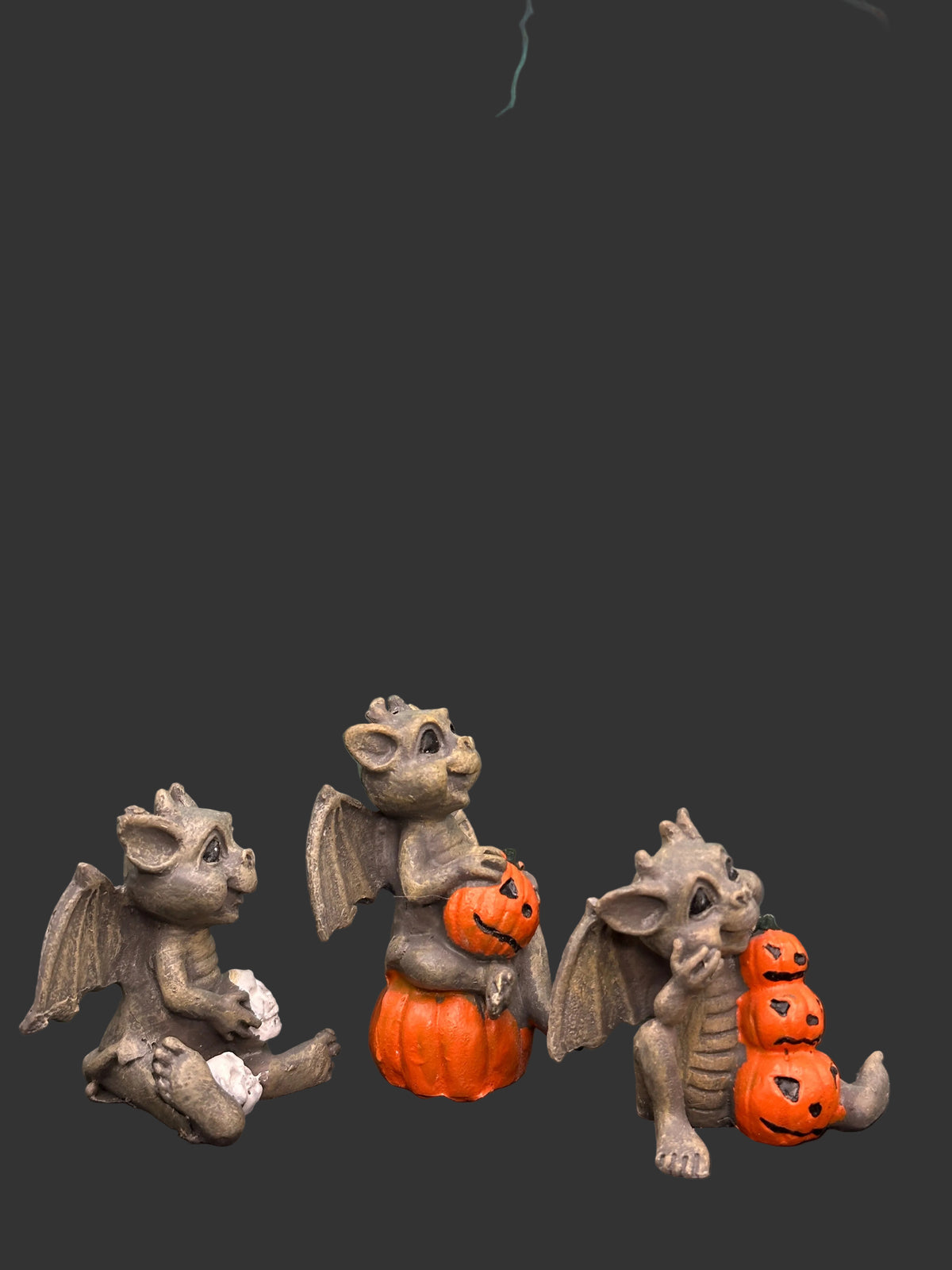 Three miniature gray stone-like gargoyle resin figurines posed on a black surface in front of a black featuring a gate, moon, trees, and orange jack-o'-lanterns.