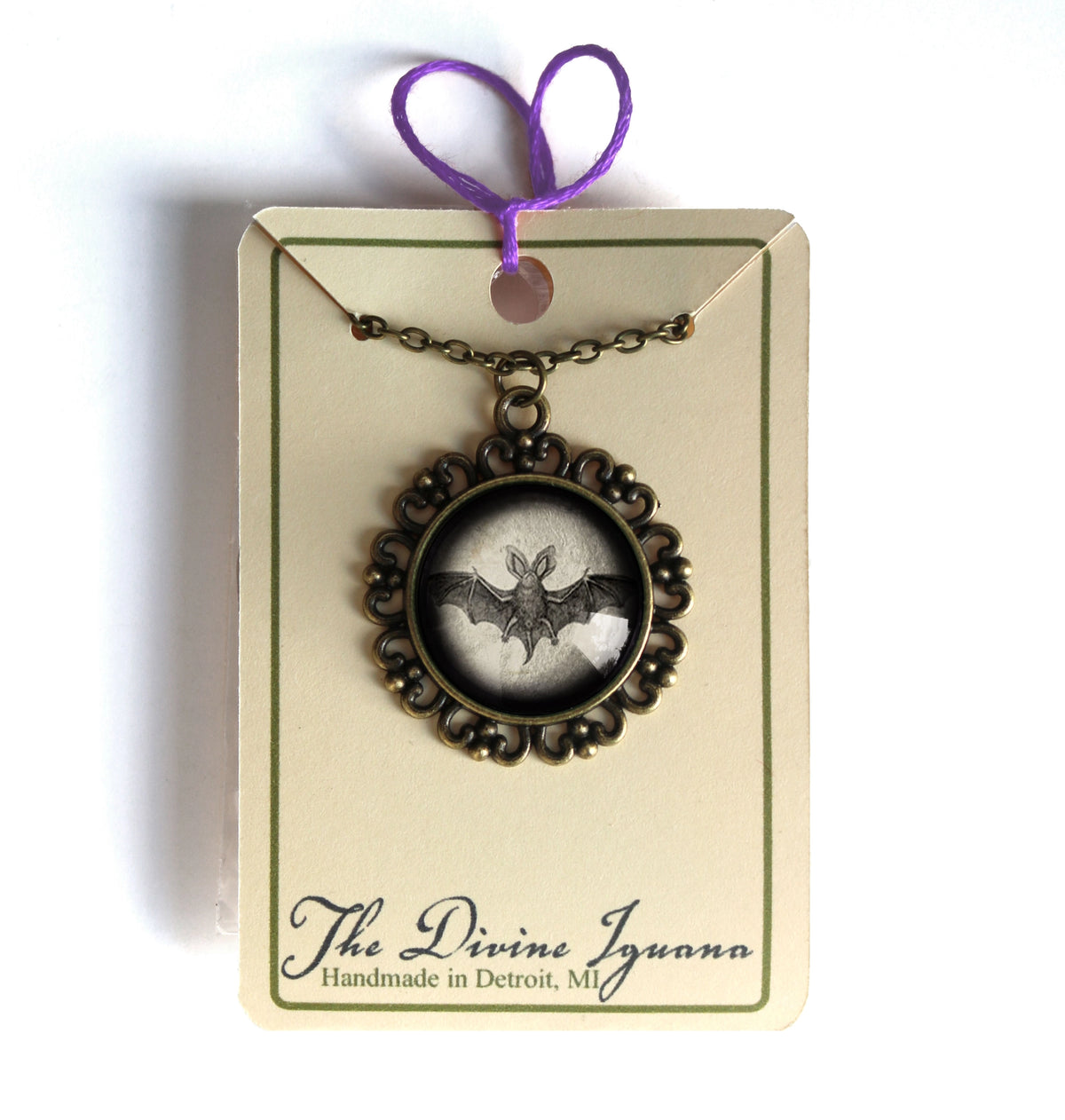 A gothic-style bat pendant necklace on a vintage bronze-toned chain, presented on a cream-colored gift card with a purple string. The card features the brand name 'The Divine Iguana' and is photographed against a plain white background.