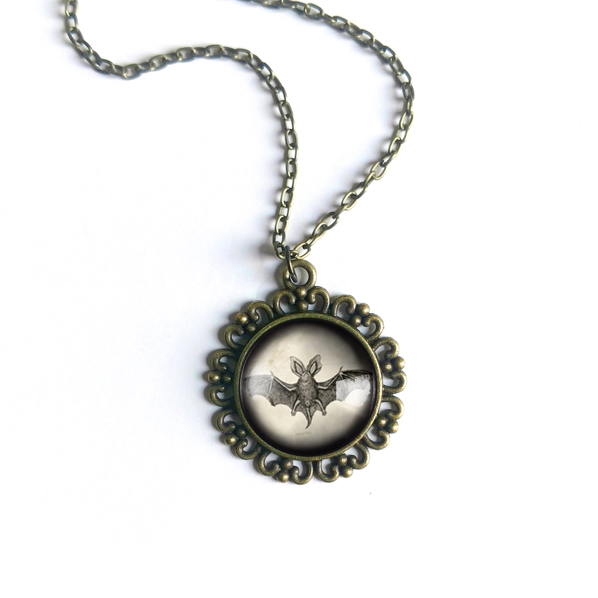 An antique bronze gothic pendant necklace featuring a bat design under a glass dome, set in an ornate filigree frame on a matching chain against a white background.