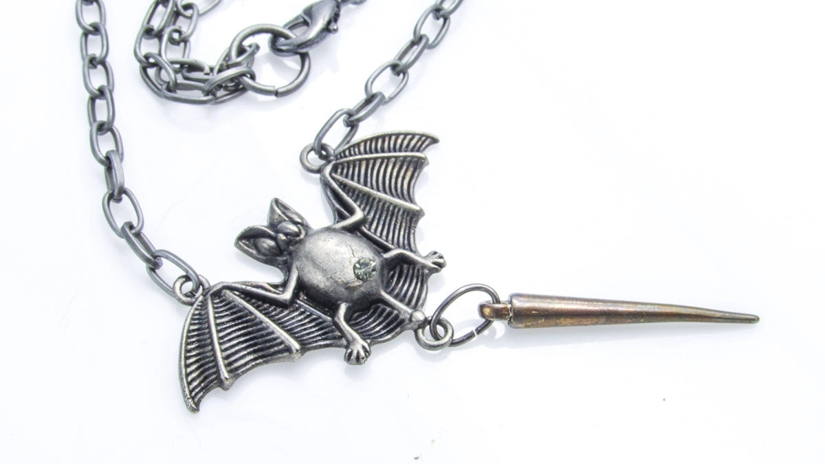 A gothic-style metal pendant necklace featuring a detailed bat charm with spread wings and a sharp, spiked tail, attached to a silver-toned link chain on a white background.