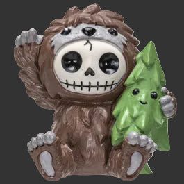 A collectible Furrybones figurine of a brown furry Bigfoot creature wearing a skull mask and holding a small green Christmas tree, set against a plain black  background.