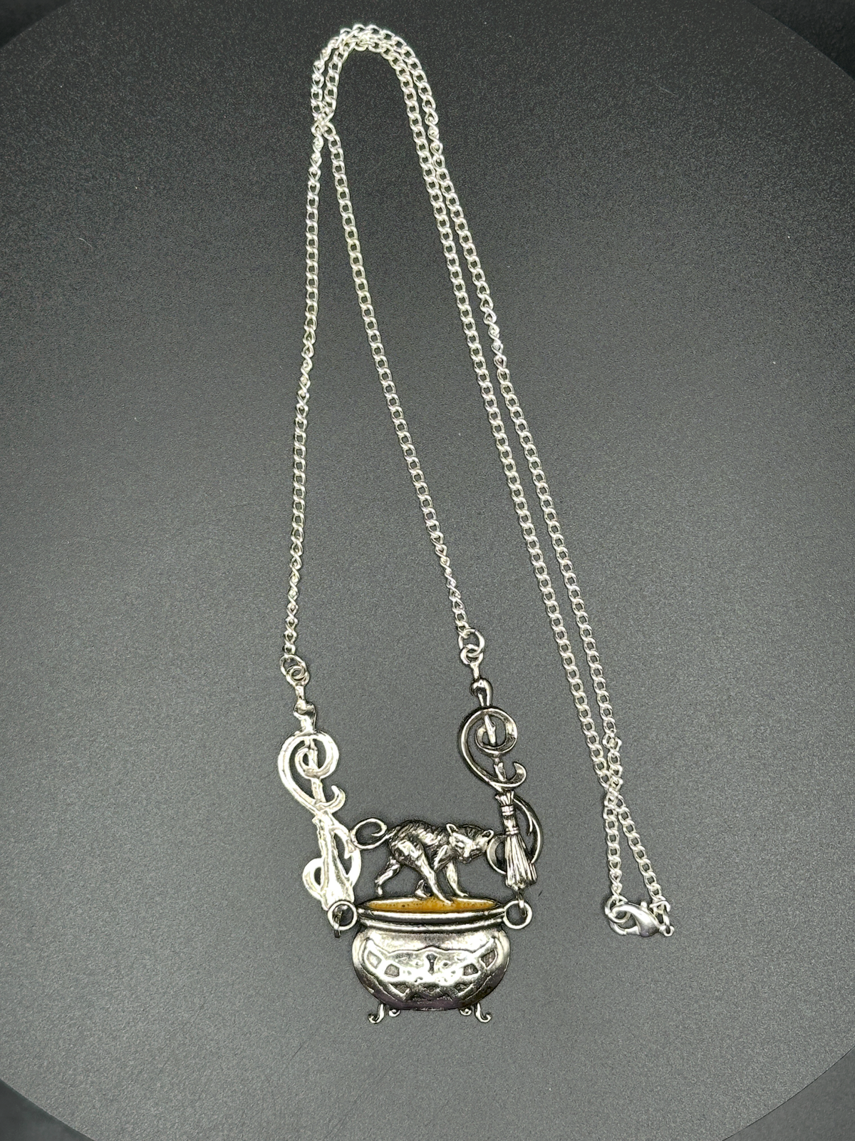 A silver-toned pendant necklace featuring a small decorative cauldron with a gold accent and musical note details, suspended from a silver chain against a dark gray background.