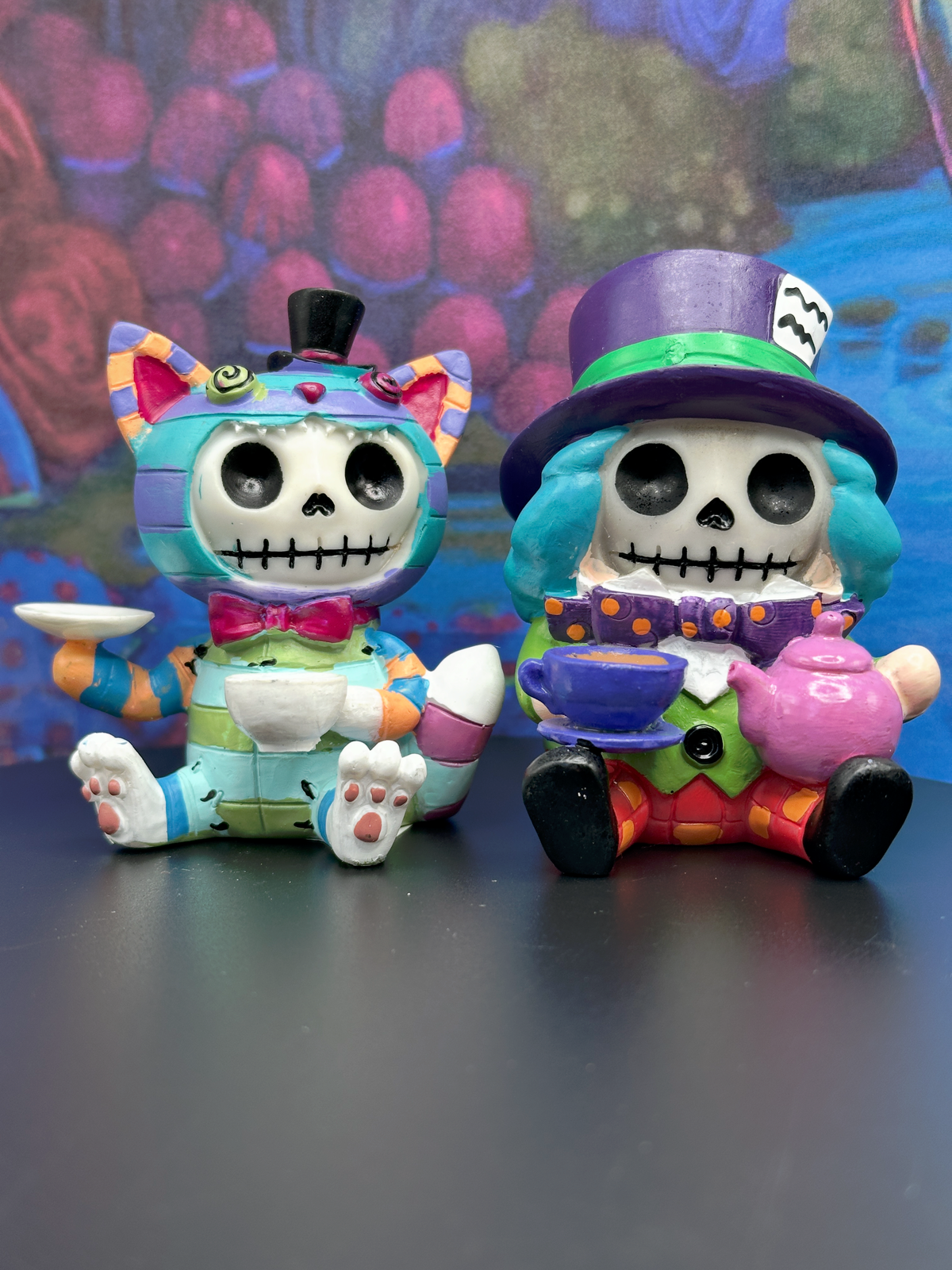 Two colorful skull-themed Furrybones Alice in Wonderland Chesire Cat and Mad Hatter figurines posed side by side on a dark surface against a painted multicolor background. The left figurine has pastel multicolor stripes, cat ears, a small black top hat, and holds a white teacup.
