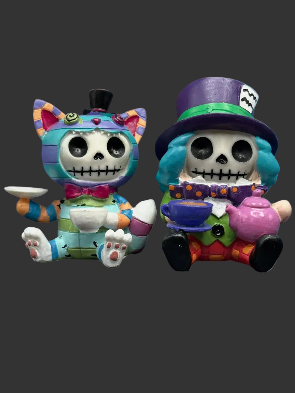 Two colorful skull-themed Alice in Wonderland collectible furrybones figurines Chesire cat and teh Mad Hatter posed side by side on a dark surface against a painted purple and blue background. The left figurine has turquoise hair, a black top hat, and a pink bow tie while holding a white teacup.