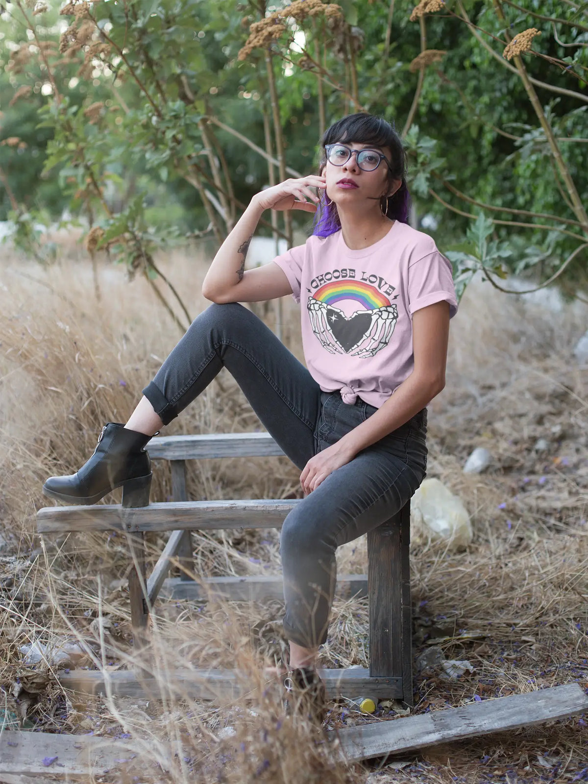 A woman wearing a pink graphic t-shirt with a rainbow and heart design, paired with black jeans and black ankle boots, sitting on a weathered wooden bench in an outdoor grassy area.