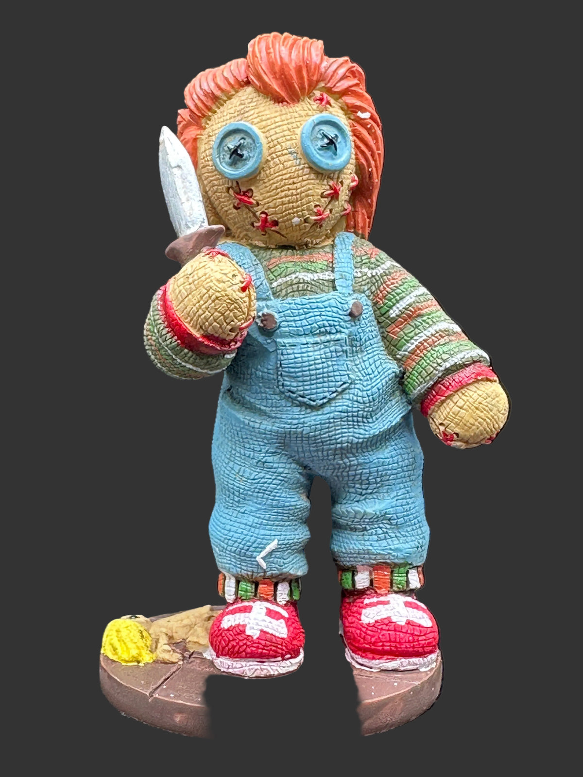 A handmade cold cast resin figurine resembling Chucky, standing on a round brown base. The doll has orange yarn hair, blue button eyes, a striped green and red shirt, blue knitted overalls, and red shoes with white crosses.