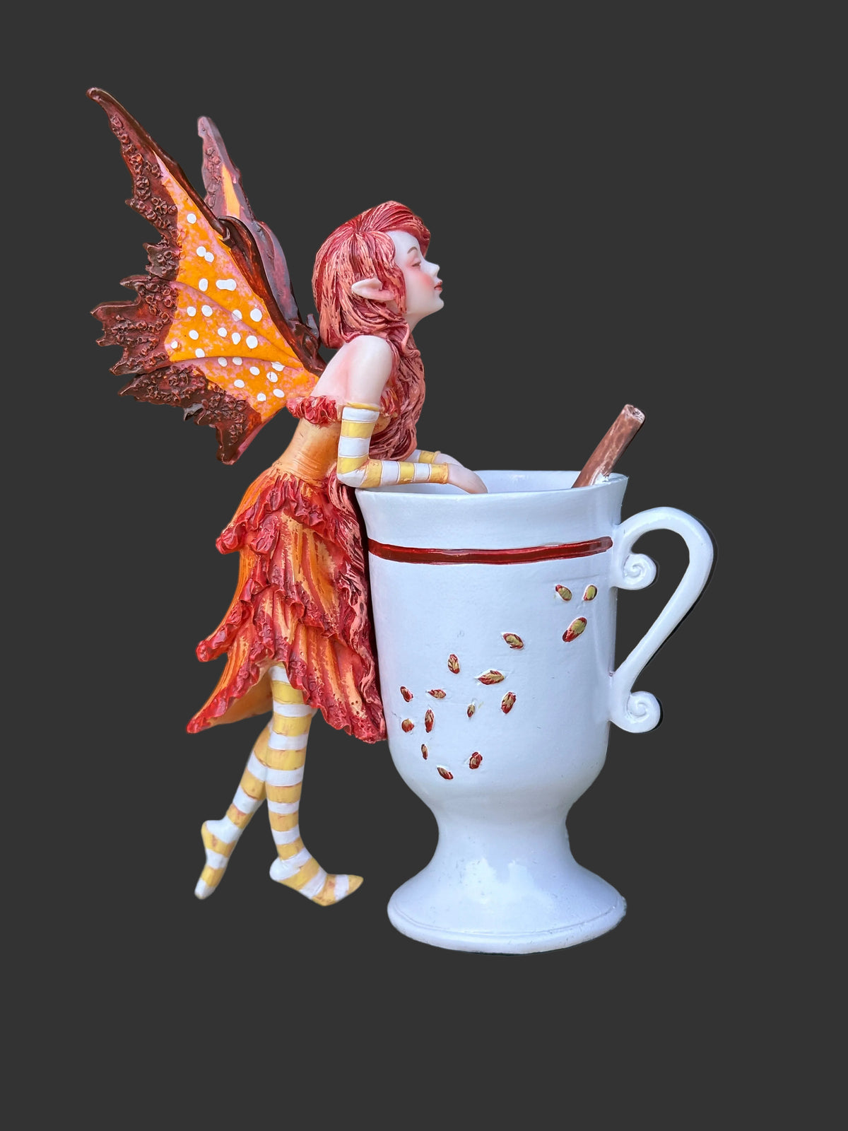 A decorative Amy Brown fairy figurine with orange wings and red hair leans against a white ceramic mug with brown speckles, placed on a stone surface with green foliage in the background.
