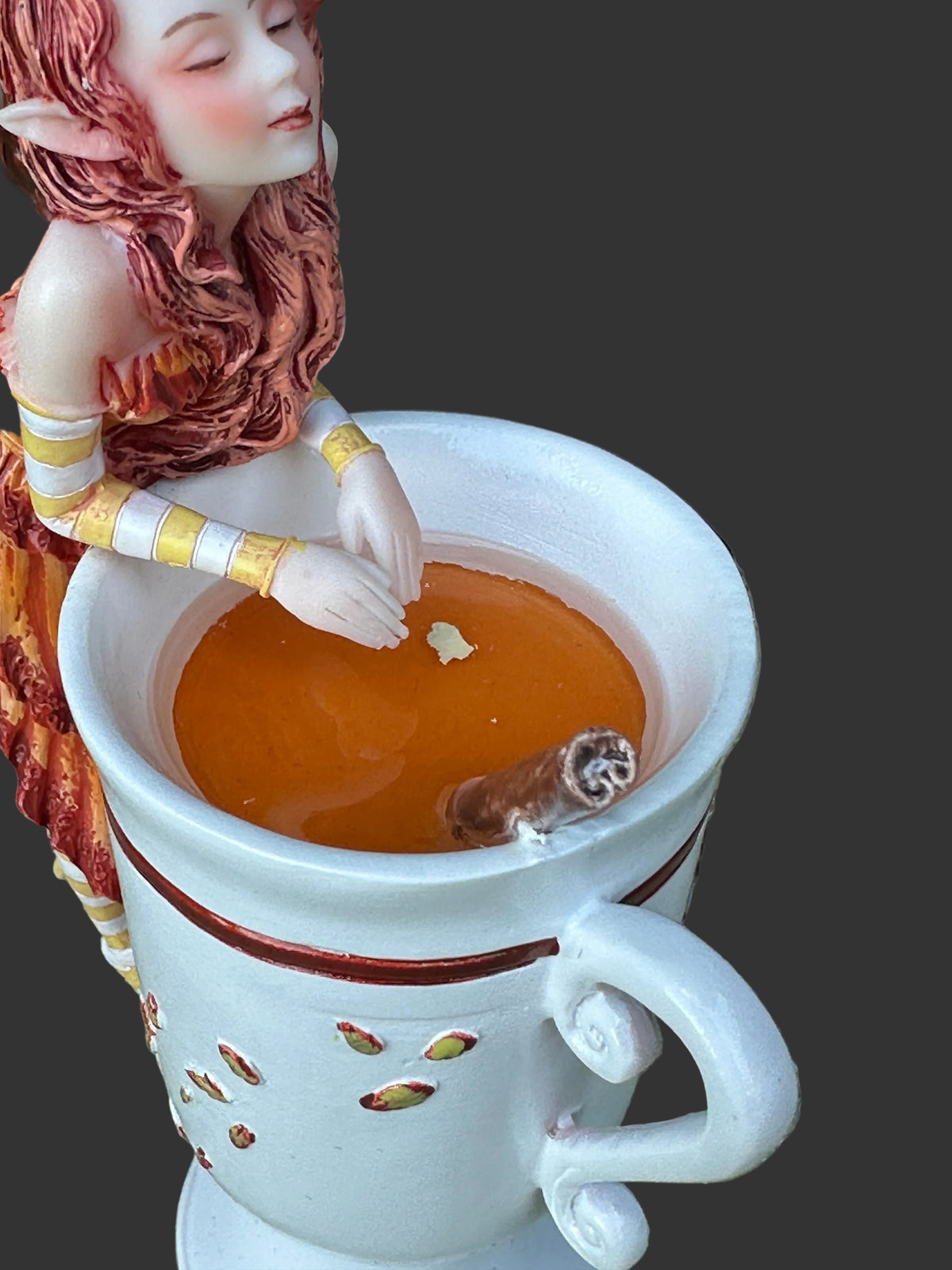 A white ceramic fairy Amy Brown mug with a handle, filled with amber-colored liquid and a cinnamon stick, set outdoors on a stone surface with a decorative elf figurine beside it.