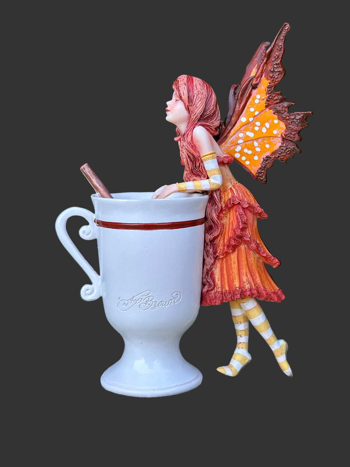A decorative Amy Brown fairy figurine in orange and red tones with patterned wings and striped legs, posed beside a white ceramic mug on a stone surface with blurred green foliage in the background.
