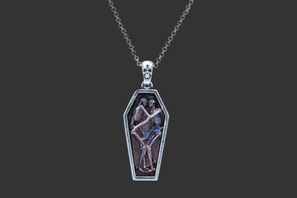 A silver coffin-shaped pendant necklace featuring a detailed illustration of two skeletons embracing, set against a dark background with pink and blue glitter accents, suspended from a silver ball chain on a white background.