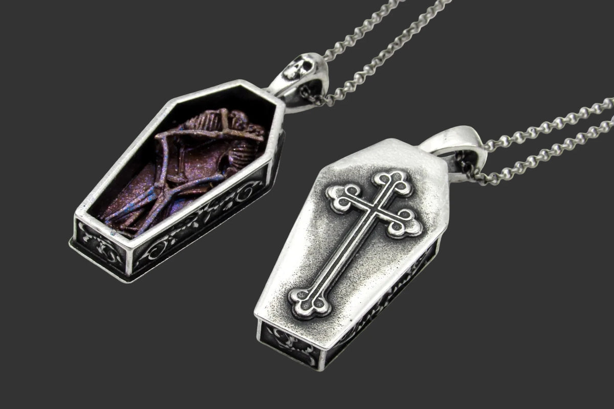 Coffin-shaped pendant necklace on silver ball chains against a white background. One pendant shows a dark purple and blue iridescent interior with a skeletal figure, and the other shows the front of the coffin, an antiqued silver cross on the lid.