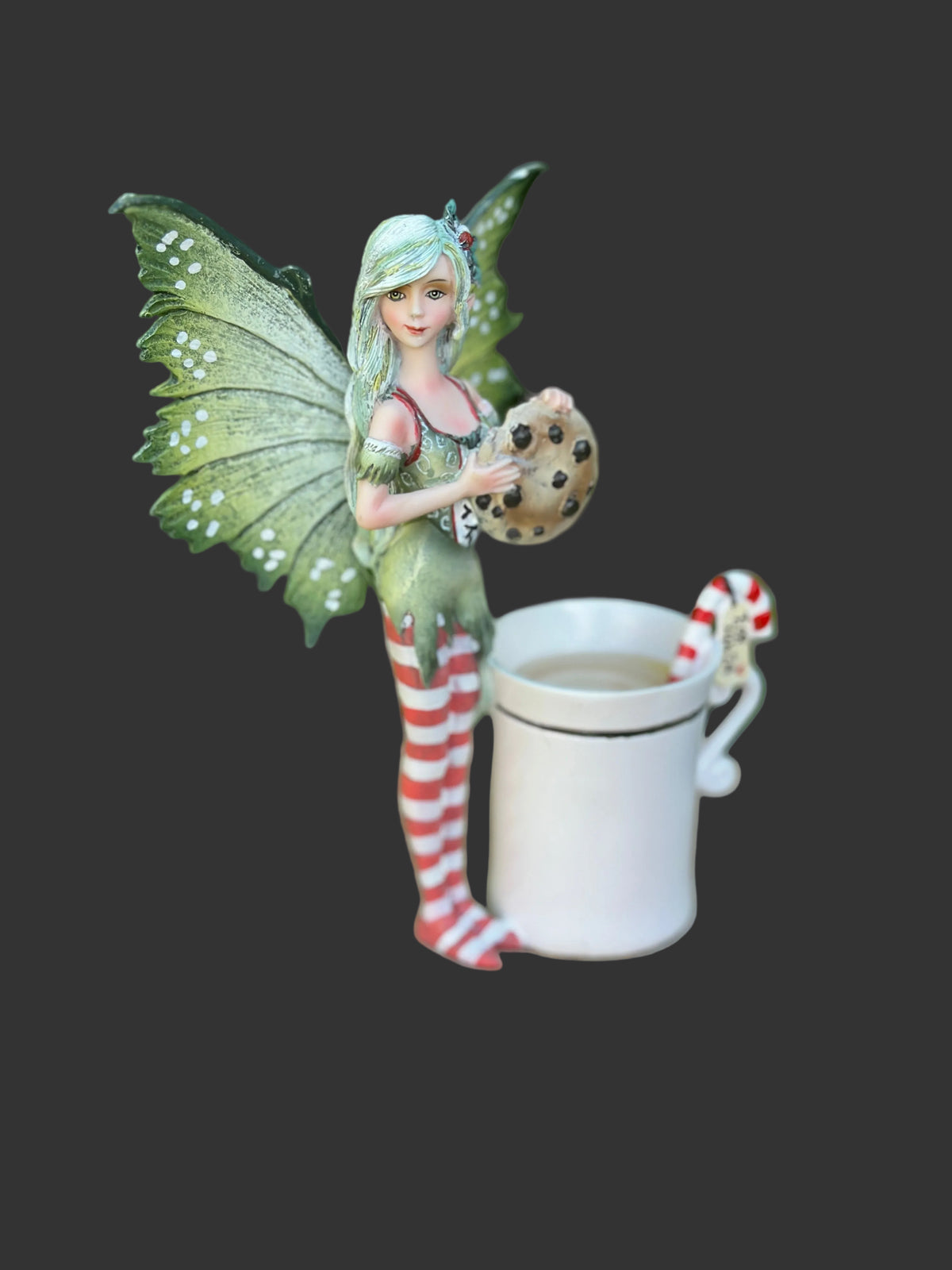 A decorative Amy Brown holiday fairy figurine with green wings and red-and-white striped stockings stands beside a white mug on a round wood slice, with a blurred green outdoor background.