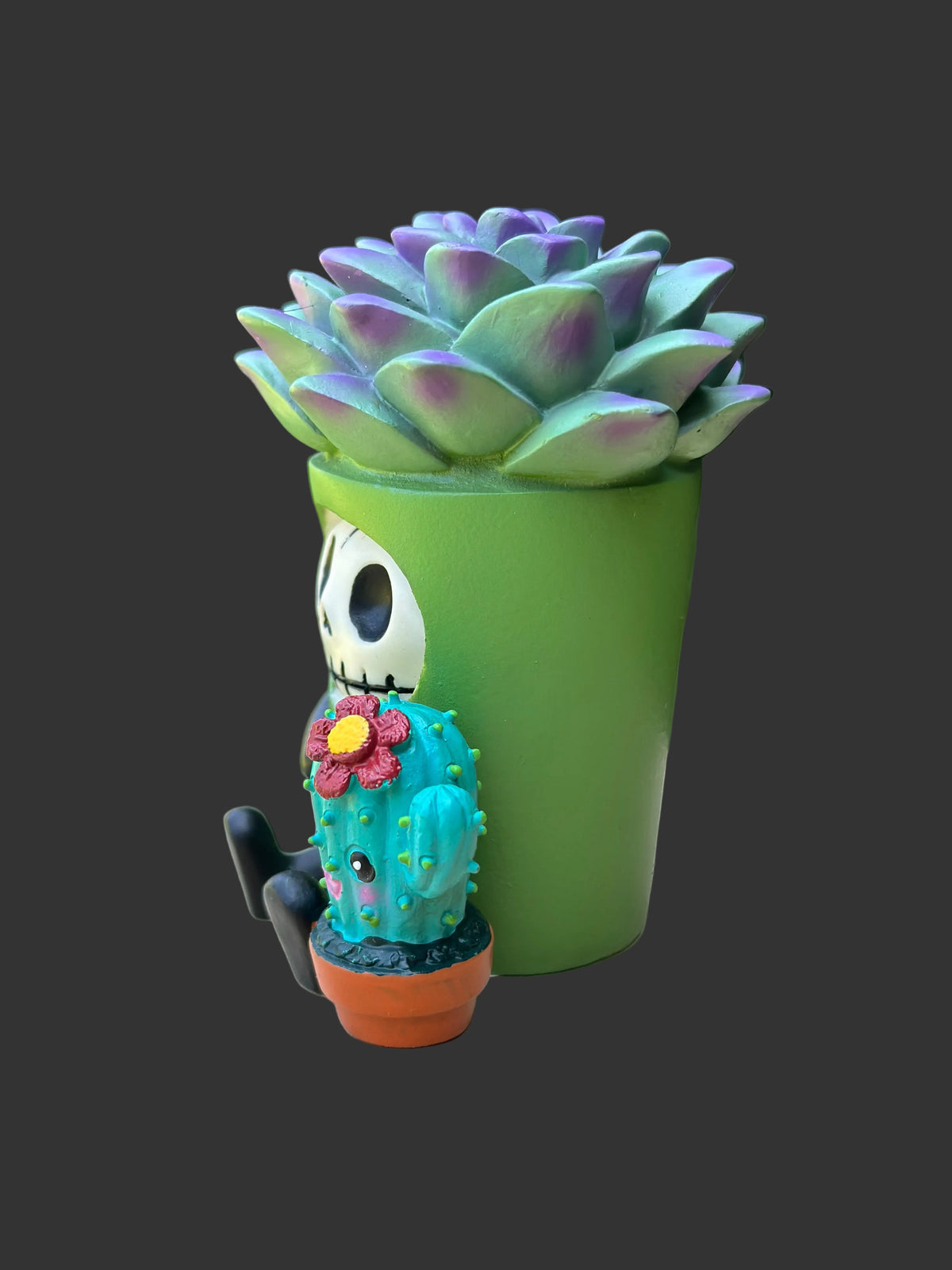 A decorative Furrybones succulent figurine and cactus garden scene with a green planter shaped like a cartoon skull face, a purple-green succulent on top, and a small turquoise cactus in a terracotta pot with a flower,