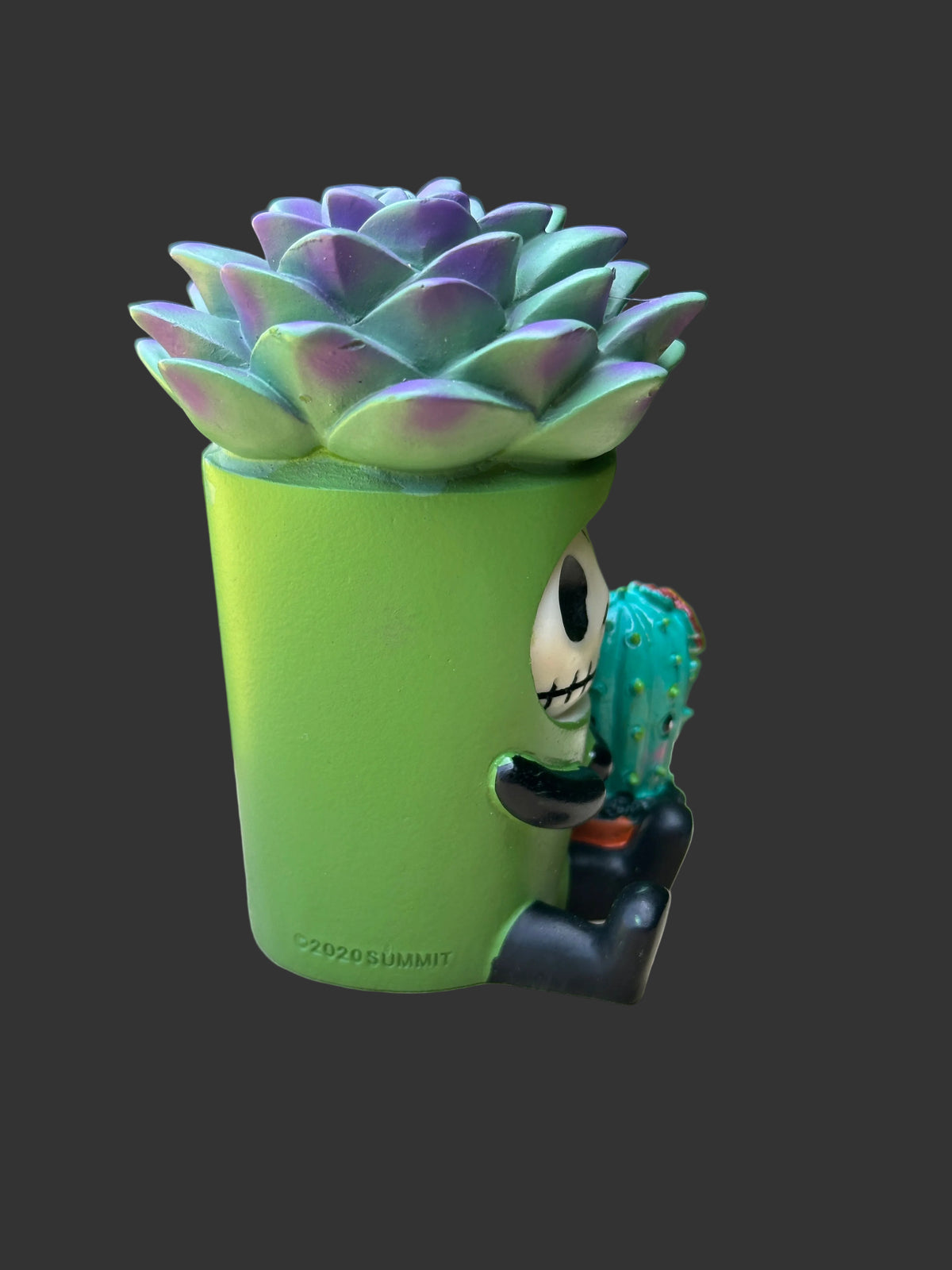 A spooky furrybones succulent figurine in a bright green cylindrical pot shaped like a mug, with a cartoon character skull peeking out from the side holding a small cactus. The succulent has iridescent green, blue, and purple tones.