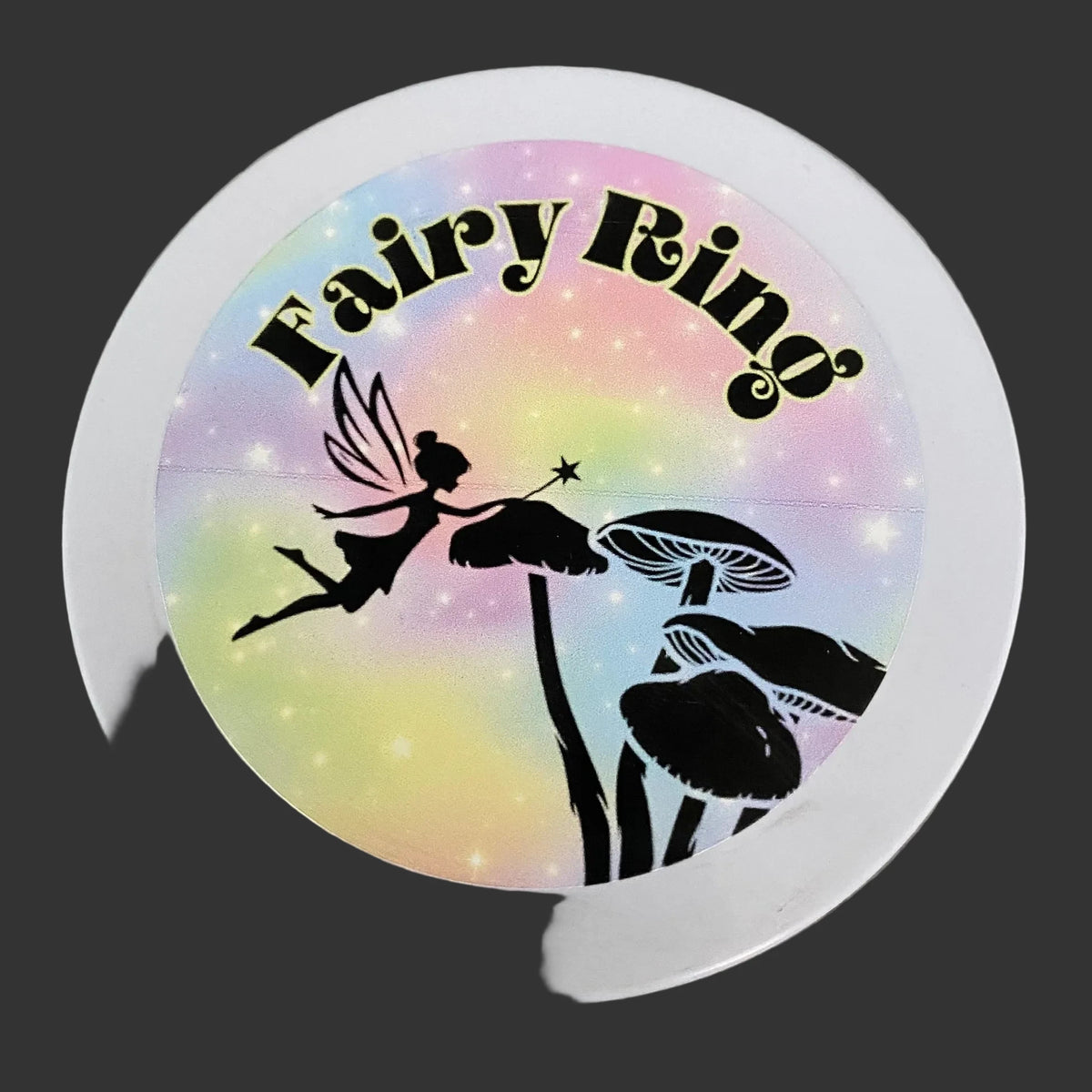 A round white ring with a colorful printed label featuring the words 'Fairy Ring' in black script over a pastel rainbow background with a black silhouette of a fairy, mushrooms, and stars.