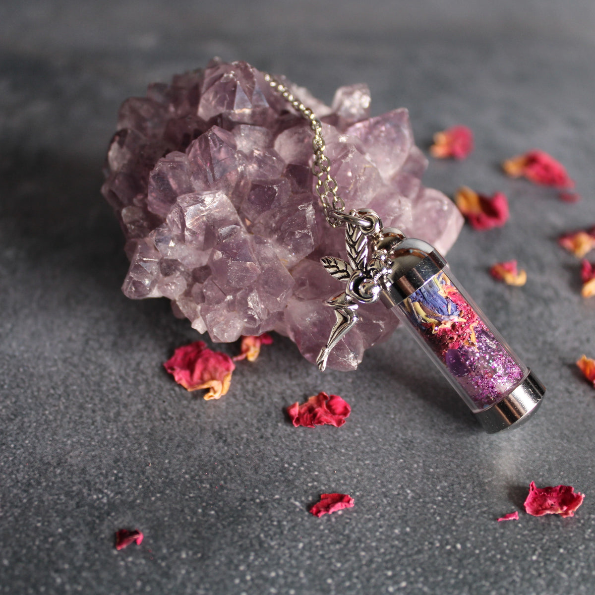 A silver fairy pendant necklace with a glass vial filled with colorful glitter and dried flower pieces, displayed on a dark gray surface next to a purple crystal cluster with scattered rose petals.