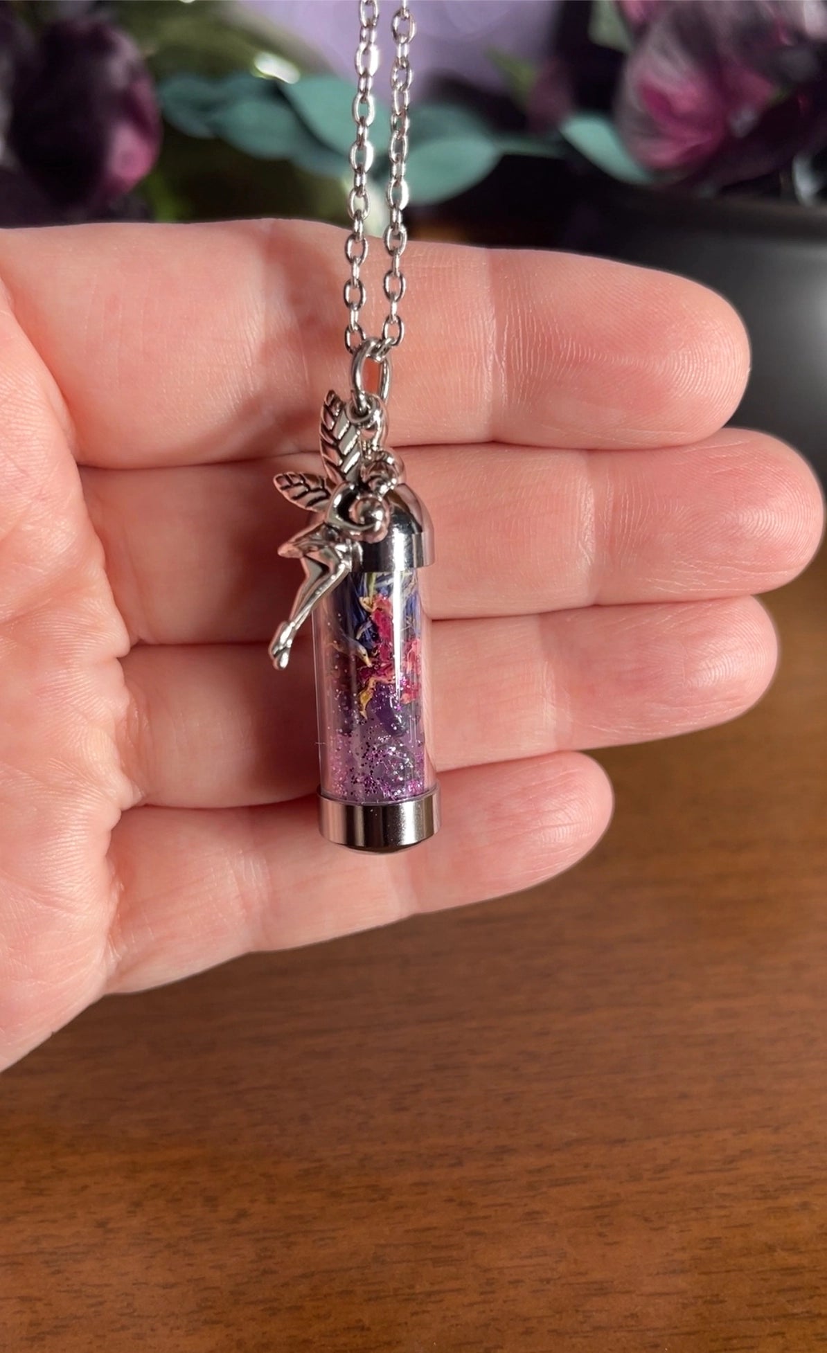 Spell Jar Necklace with a pink and silver fairy pendant held in a hand on a wooden surface.