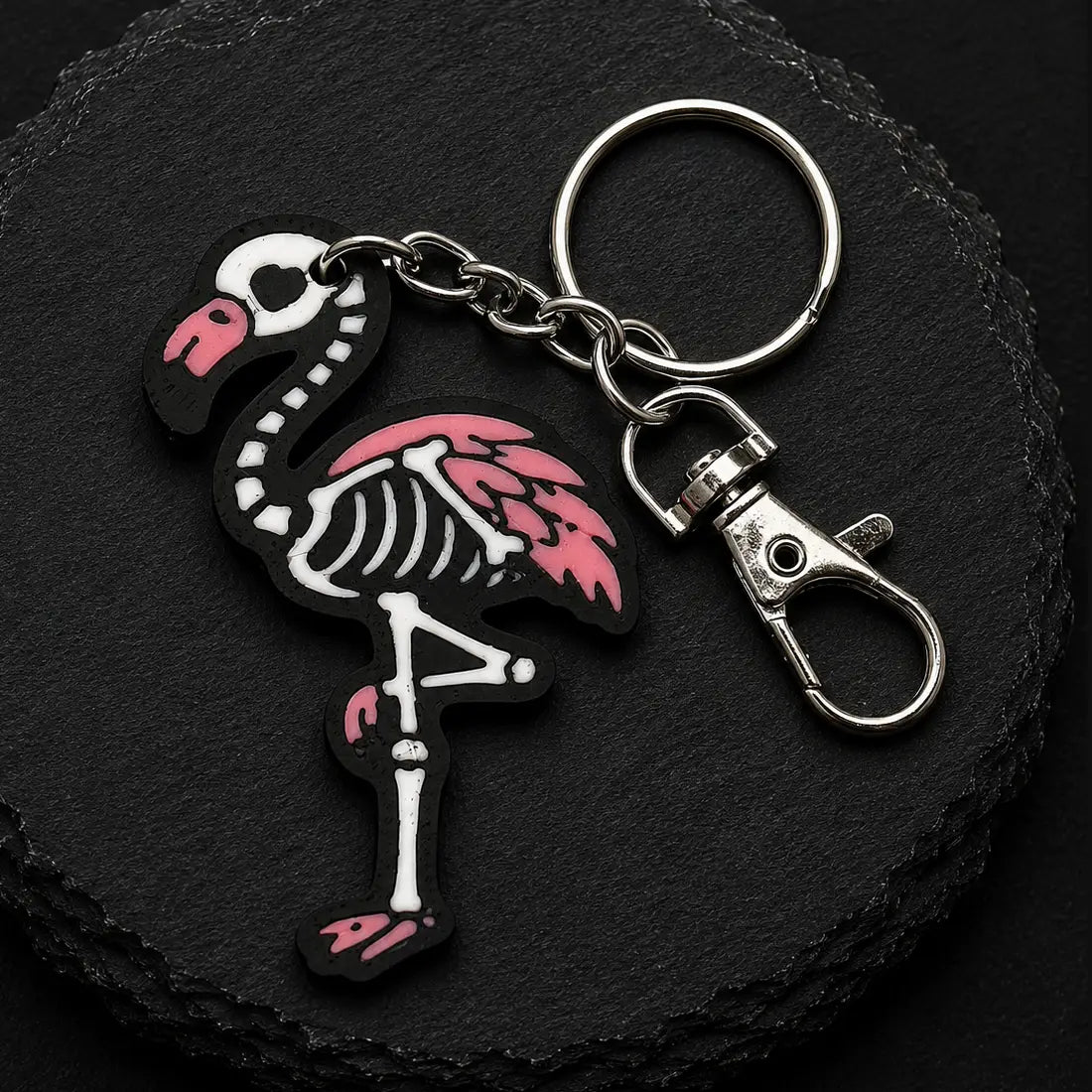 A flamingo skeleton keychain with a pink and white design, attached to a silver metal split ring and lobster clasp, displayed on a black slate background.