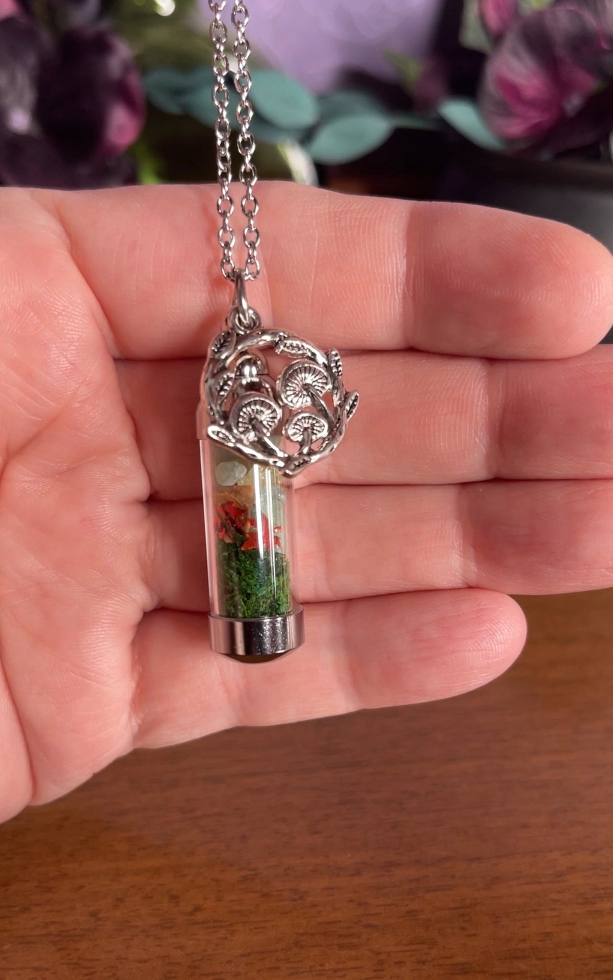 A silver-toned necklace with a cylindrical pendant held in a hand. The pendant features a clear glass vial filled with green moss and small red flower pieces, topped with an ornate silver filigree cap.