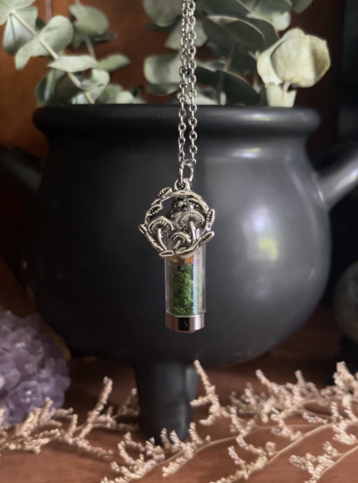 Spell Jar Forest Necklace with a decorative mushroom pendant in front of a black pot and plants.
