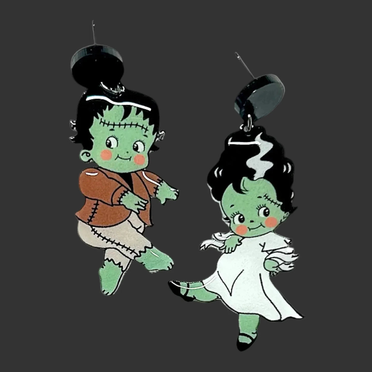 A pair of dangling earrings featuring cartoon Frankenstein's monster and a bride Elizabeth  in a white dress, shown against a blurred black-and-white background.