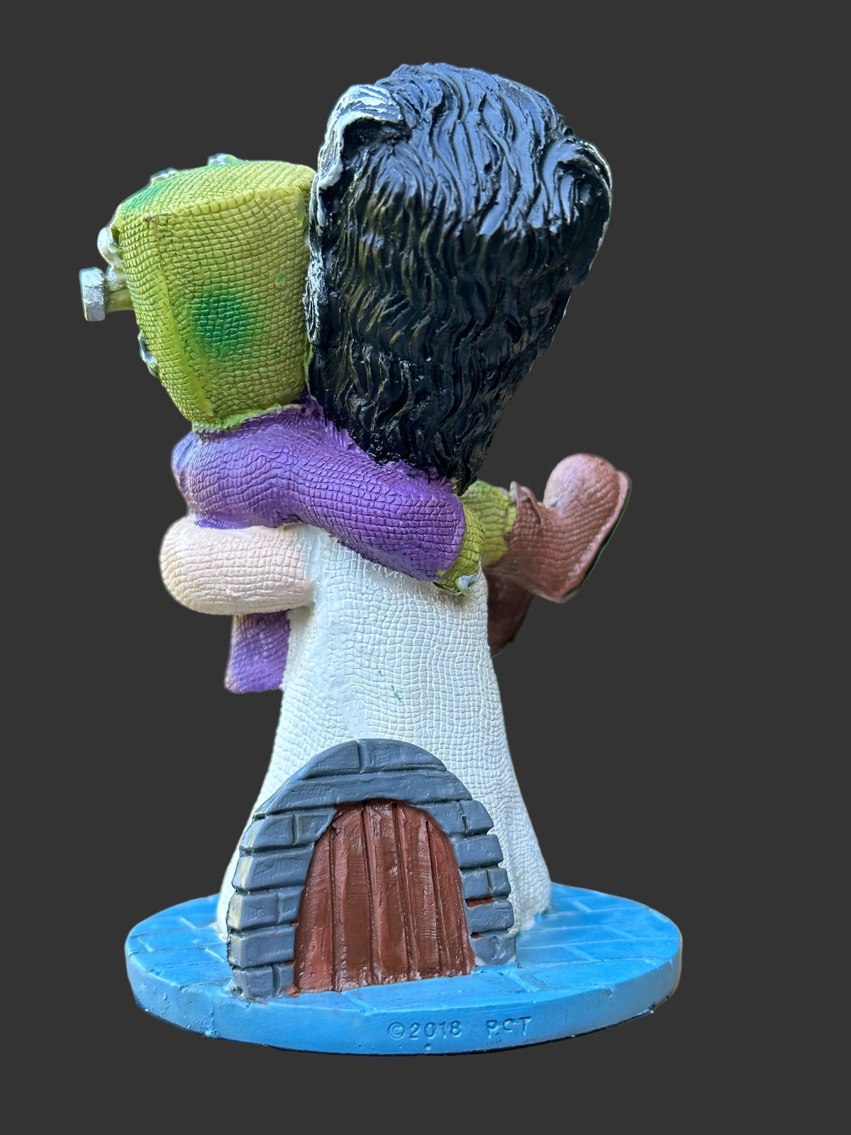 A small decorative figurine of a hugging couple, Frankenstein and Elizabeth one figure in a white robe and blue base and the other in a green top and purple scarf, displayed on a wooden surface with a black background.