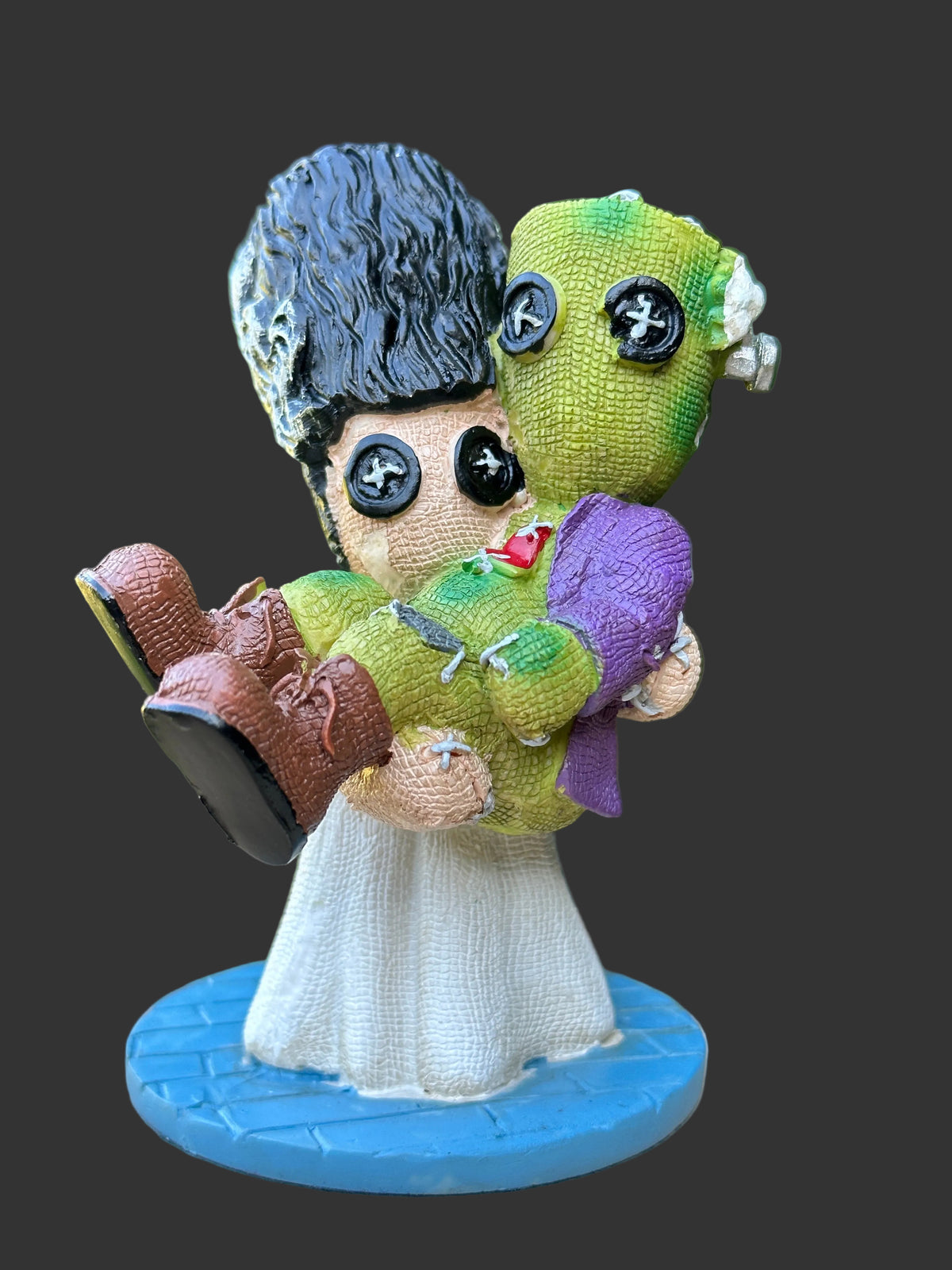 A handmade cold cast resin doll figurine Frankenstein's bride, Elizabath holding Frankenstein,  displayed on a wooden surface with a black background. The doll features a black hair bun, a white dress, a green bodysuit, and a purple blanket, with realistic button eyes and stitched details.