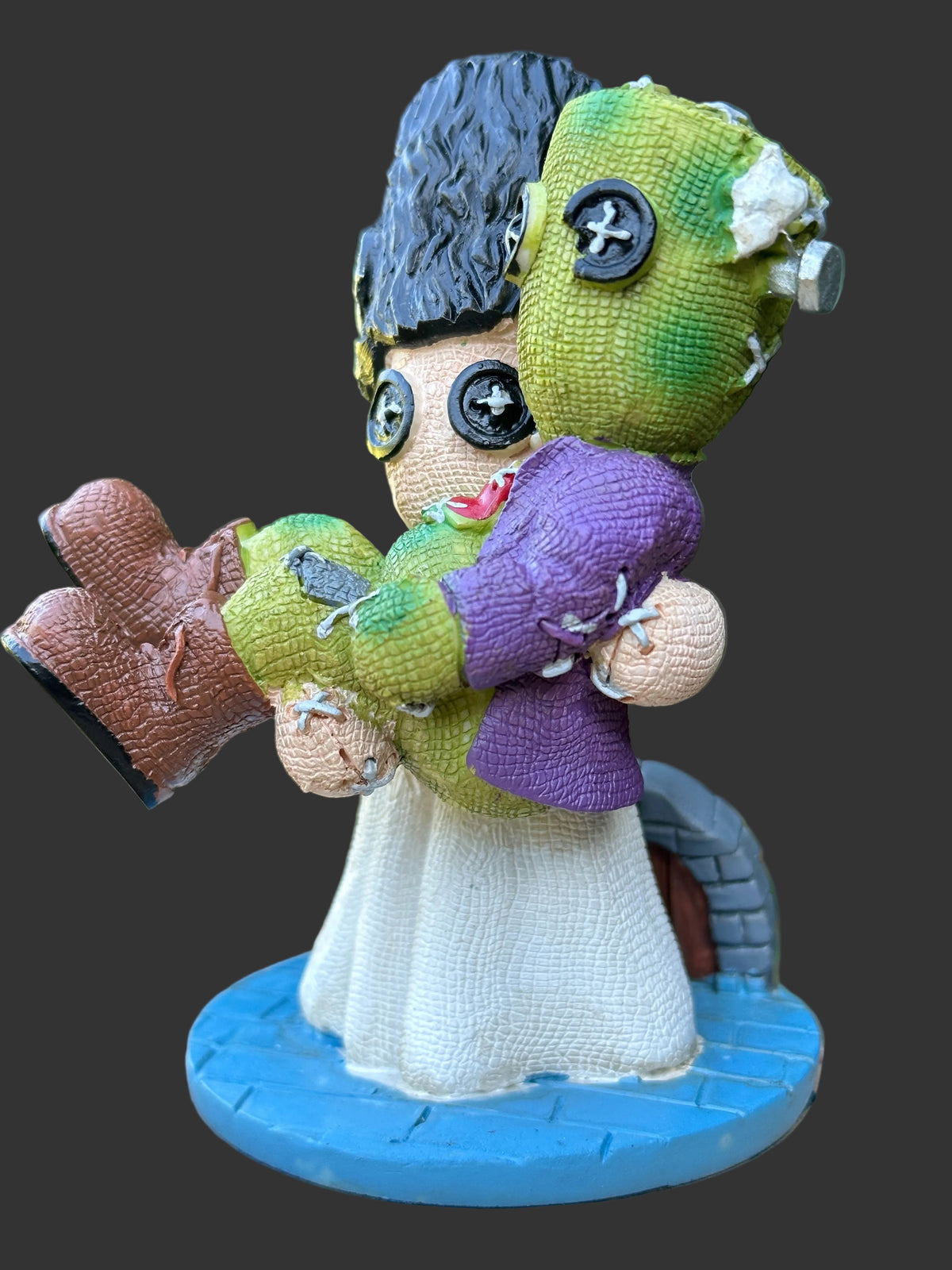 A handmade cold cast resin figurine of Frankenstein and his bride holding a each other, standing on a blue circular base. The larger doll has black yarn hair, a beige textured body, a white dress, and is holding a smaller doll with green and purple yarn clothing.