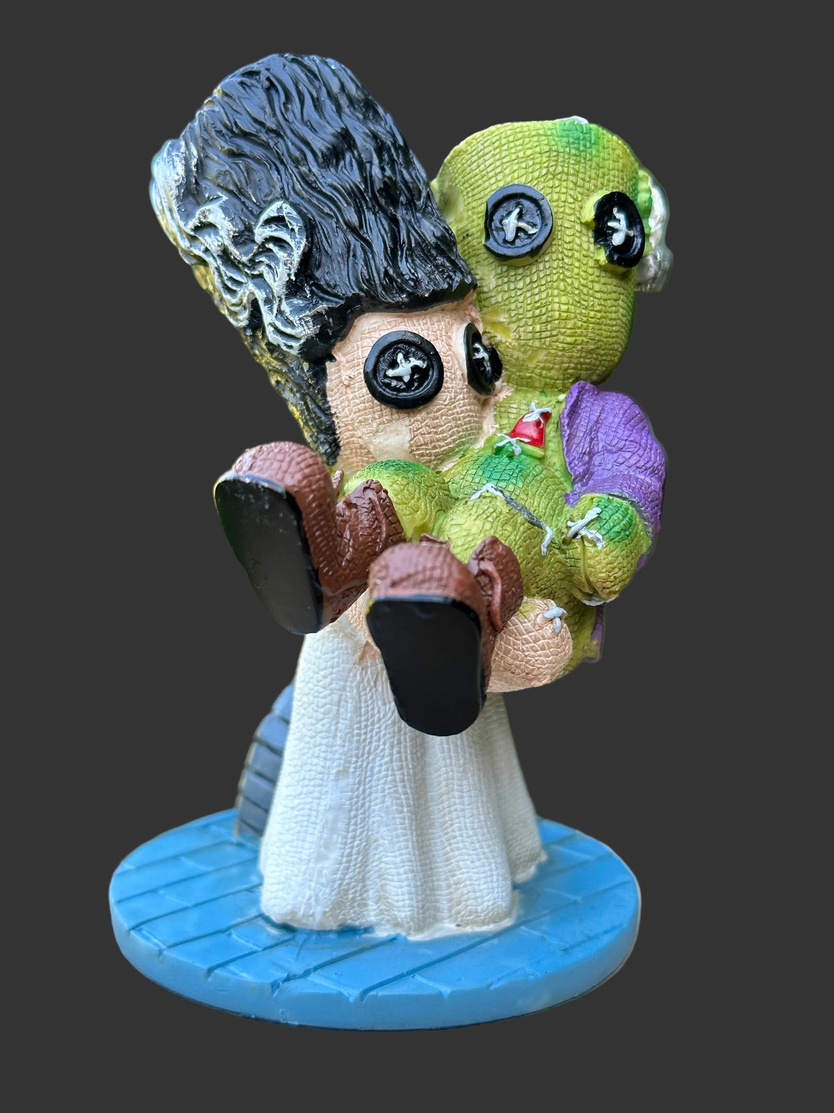 A handmade voodoo type doll figurine depicting a Frankenstein and his bride, a woman in a white dress holding a green stitched doll with button eyes, standing on a blue circular base.
